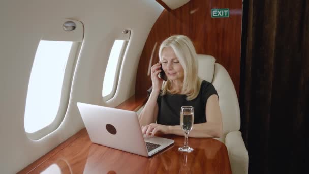 Laptop bilgisayarında çalışan ve özel uçakta seyahat ederken cep telefonuyla konuşan yetişkin bir milyoner kadın. Özel jetteki iş kadını ya da bilgisayarlı birinci sınıf iş kadını. — Stok video