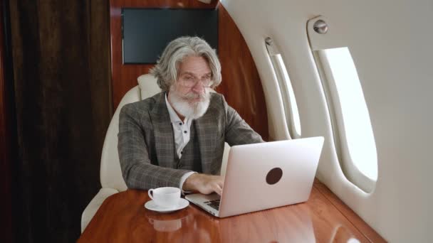 Excited mature male looks at laptop and rejoices in victory while flying airplane. Happy gray-haired rich man working at computer and celebrating business success while traveling in private jet plane — Stock Video
