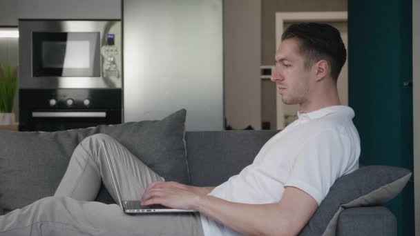 Freelancer jovem usando dispositivo laptop enquanto deitado no sofá em casa, retrato de homem trabalhando distantemente no computador no apartamento — Vídeo de Stock