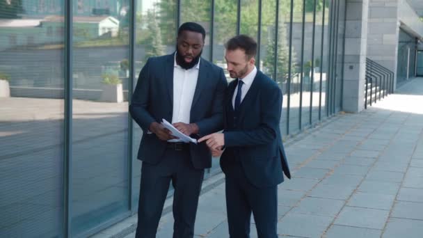 Resmi takım elbiseli iki farklı iş adamı ellerinde kağıt parçalarıyla iş hakkında konuşuyor ve tartışıyorlar. Şehir merkezinde ofis binasının yanında duruyorlar. Erkek girişimciler dışarıda konuşuyor.. — Stok video