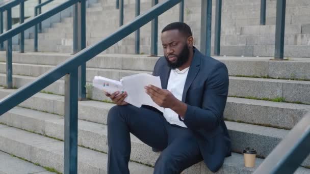 L'uomo afroamericano barbuto guarda attraverso documenti cartacei con grafici aziendali mentre si siede sui gradini vicino al centro dell'ufficio. — Video Stock
