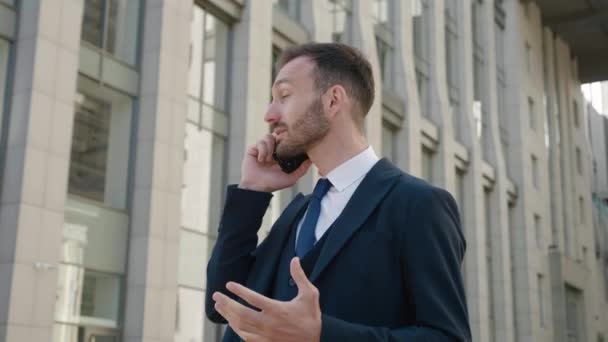 Przystojny brodaty biznesmen w formalnym garniturze emocjonalnie mówiący przez telefon komórkowy stojąc na zewnątrz w centrum, w pobliżu biurowców. — Wideo stockowe