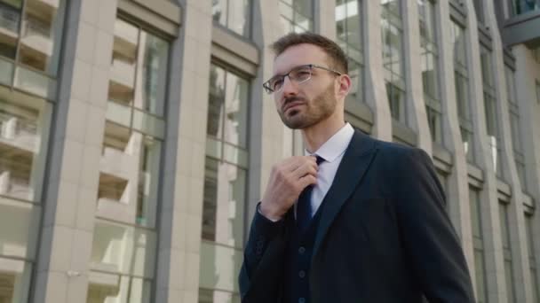 Portré jóképű üzletember szemüvegben, aki az irodaház közelében sétál. Egy öltönyös férfi néz félre a szabadban a városban. — Stock videók