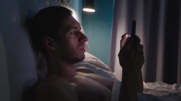 Close-up van een man die 's nachts op een bed in de slaapkamer ligt en een smartphone gebruikt. Jonge man surfen op het internet met een mobiele telefoon voor het slapen gaan. — Stockvideo