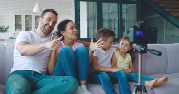 Γυναίκα κάθεται δίπλα στην οικογένειά της στον καναπέ, κάνοντας βίντεο blog σε επαγγελματική βιντεοκάμερα, ενεργά χειρονομεί, αναφερόμενη στον άντρα και τα παιδιά της, περιγράφει το νέο δωμάτιο με την εγγραφή βίντεο — Αρχείο Βίντεο