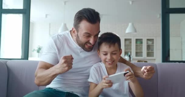 4K video van verbaasd kind jongen actief spelen van computerspelletjes op zijn mobiele telefoon en zijn jonge vader actief ondersteunen hem in het spel — Stockvideo
