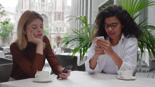 Dwie znudzone, zróżnicowane przyjaciółki przeglądają internet na smartfonach w kawiarni na świeżym powietrzu. — Wideo stockowe