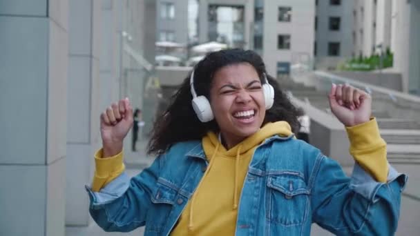 Souriante jeune fille afro-américaine qui aime écouter de la musique en ville à l'extérieur. Heureuse femme métissée portant des écouteurs dansant seule dans la rue. — Video