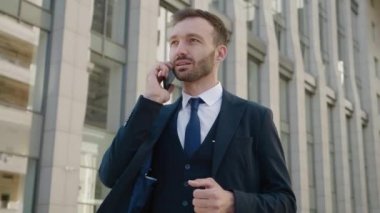 Resmi takım elbiseli yakışıklı iş adamı şehir merkezindeki ofis binalarının yanında beklerken cep telefonuyla konuşuyor.