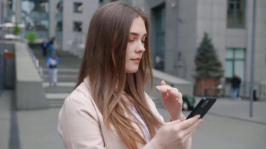 Endişeli iş kadını akıllı telefonuyla kötü haber okurken iş yerinin dışında dikiliyor. Ofis binasının dışında elinde cep telefonu tutan genç bir kadın şok oldu.