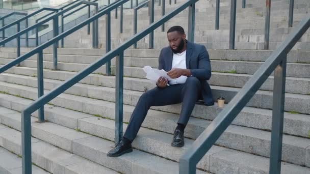 Одягнений афроамериканець переглядає паперові документи з діловими картами, сидячи на сходах біля офісного центру.. — стокове відео