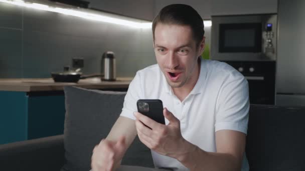 Surpreendido jovem vencedor segurar smartphone ler boas notícias espantado com celular on-line aposta lance jogo vitória em casa. Happy male look at cell phone screen overjoyed by victory success while sitting on sofa. — Vídeo de Stock