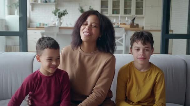 Family portrait of a cute African American young mother enjoying time with her two adorable toddlers, smiling, communicating sitting on the living room sofa, looking at camera on the kitchen backdrop. — Stock Video