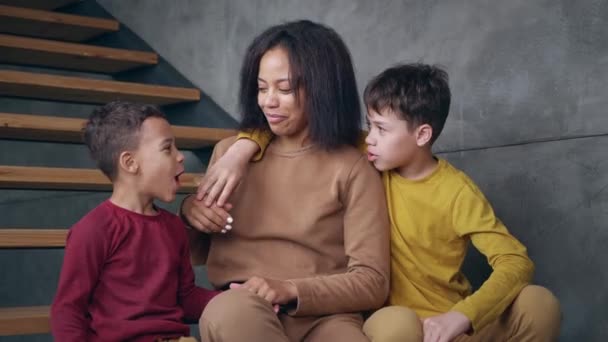 Young African American woman, sitting on the home stairs between her adorable sons, stroking the younger on the head. The older hugging his mother. Love, tenderness and happy motherhood concept — Stock Video