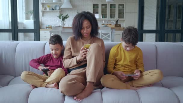 Joven mujer afroamericana sentada junto a sus hijos en el sofá de la sala de estar. Los tres se centran en divertirse con sus aparatos digitales. Familia feliz de tres vínculos descansando en casa — Vídeo de stock