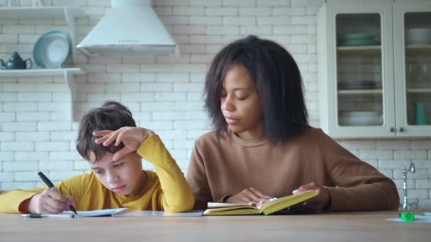 Young woman helps her son with homework. Caring mom checks her childs school tasks . Boy writing a dictation according to his mothers guidance at home. Concept of back to school, Knowledge, study. — Stock Video