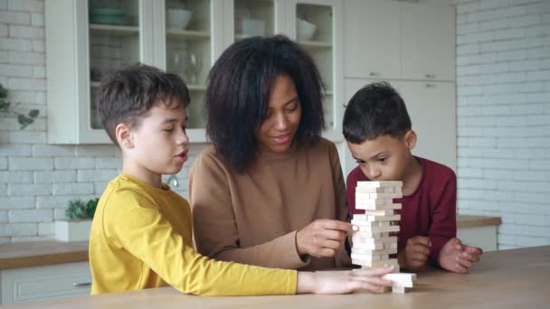 Афроамериканская мама с детьми, играющими в настольные игры, смеялась после падения деревянной башни из высоких игрушек. Семья проводит время вместе дома. Наслаждение образовательными играми во время блокировки или выходных — стоковое видео