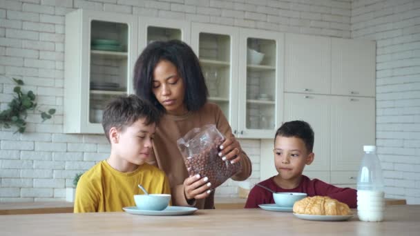 Madre afroamericana vertiendo copos de chocolate de cereales en los cuencos. Niños desayunando sentados a la mesa en la cocina. Botella con leche y croissants sobre la mesa en primer plano — Vídeos de Stock