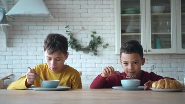 İki güzel anaokulu öğrencisi kahvaltı için lezzetli çikolata gevreğinin tadını çıkarırken mutfak masasında oturuyorlar. Sevimli çocuklar sağlıklı bir kahvaltı yaparken iletişim kuruyorlar.. — Stok video