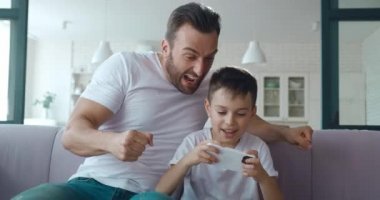 4K video portresi, akıllı telefonuyla bilgisayar oyunu oynayan yakışıklı, heyecanlı bir okul çocuğunun ve genç babasının oyunda aktif olarak onu desteklemesi.