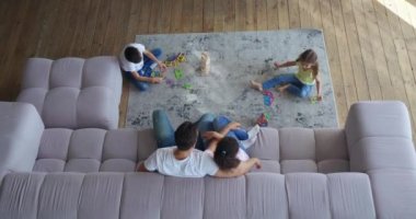 Kocasının karısına kanepede sarılıp çocuklarının oturma odasında halıda masa oyunları oynamasına hayranlık duyması. 4 K video kaydı. Kapalı alan aile eğlencesi kavramları