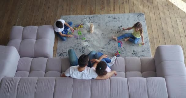 Vista superior del marido abrazando a su esposa sentada en el sofá y admirando a sus hijos jugando juegos de mesa en la alfombra en la sala de estar. 4 K Vídeo. Conceptos de ocio familiar en interiores — Vídeo de stock