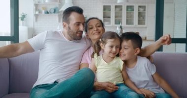 Gülümseyen erkek kardeş, küçük kız kardeşine yaslanıyor. Güzel bir ailenin portresi, kanepede yan yana dinleniyor. Yakışıklı, olgun bir baba ailesine sarılıyor. 4K video