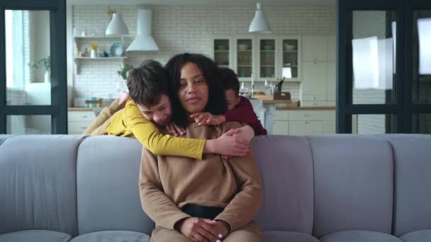 Afroameričanka, matka dvou dětí sedících doma na gauči, se dívá do kamery, zatímco její milí synové vyběhnou z kuchyně a něžně ji objímají. Raduje se a láskyplně se usmívá — Stock video