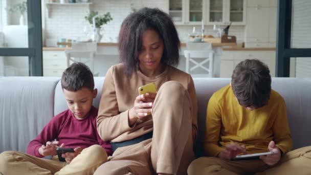 Foto close-up keluarga bersenang-senang dengan gadget digital mereka. anak laki-laki lucu melihat tablet sambil duduk di sofa sementara ibu dan adik laki-lakinya ingin tahu melihat layar sentuhannya — Stok Video