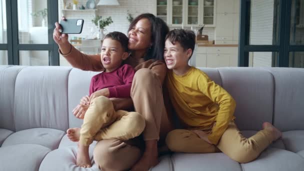 Femme africaine tenant un téléphone intelligent dans le bras tendu, étreignant ses enfants fils assis sur le canapé du salon, regardant la caméra Web, souriant, saluant en ligne par webcam faisant un vidéocall via application. — Video