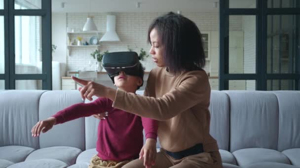 Wanita muda Afrika-Amerika, ibu bersenang-senang melihat anaknya, mengenakan kacamata virtual reality. Konsep VR. Anak laki-laki yang mengagumkan dan gelisah secara aktif bergerak selama pengalaman virtual reality. — Stok Video