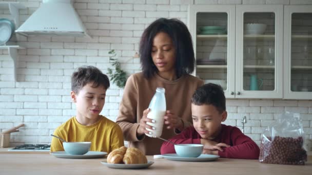 Meninos sentam-se à mesa da cozinha, à espera do pequeno-almoço. Jovem mãe derrama leite em tigelas e polvilha flocos de chocolate de cereais para seus filhos, preparando delicioso café da manhã saudável pela manhã — Vídeo de Stock