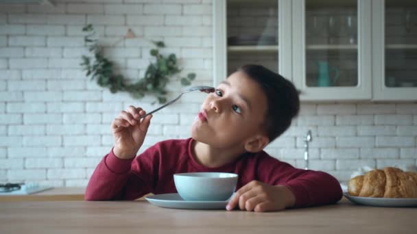 Um menino pré-escolar de raça mista senta-se em um prato de flocos de cereais de chocolate na mesa da cozinha, relutantemente come seu café da manhã, joga, examina uma colher com cereal preso a ela, se recusa a comer. Instrução — Vídeo de Stock