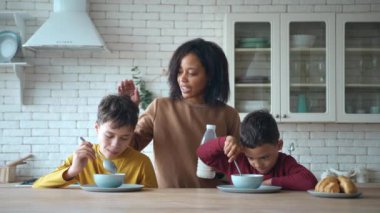 Tatlı çocuklar kahvaltıda çikolata parçacıklarıyla masada oturuyorlar. Çekici Afro-Amerikalı anne ellerinde bir şişe sütle arkalarında duruyor ve en büyük oğlanı kaşıkla oynaması için ikna ediyor.