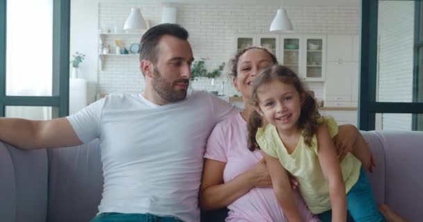 Szczęśliwa młoda para siedzi na kanapie i cute baby girl dołącza do nich, przytula matkę i ojca. Nagranie 4K o szczęśliwych związkach rodzinnych. Miłość, czułość i koncepcja czułości. — Wideo stockowe