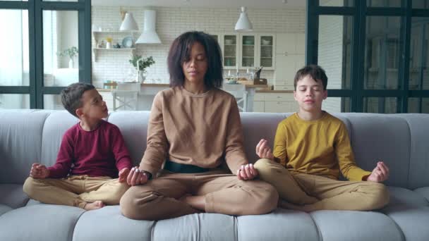 Mujer serena afroamericana sentada en el sofá en posición de loto entre sus hijos, meditando, relajándose en condiciones de calma. Familia disfrutando la meditación. Yoga, meditación, concepto de estilo de vida saludable — Vídeo de stock