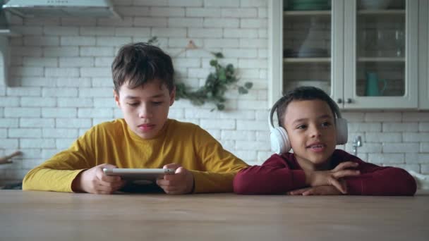 Anak laki-laki ras campuran duduk berdekatan satu sama lain di meja di restoran rumah, bersenang-senang menggunakan tablet digital, adik laki-laki aktif bergerak mendengarkan musik di headphone. Hari bebas di rumah — Stok Video