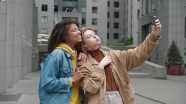 İki farklı kadın arkadaş cep telefonlarıyla selfie çekiyorlar, gülümsüyorlar ve şehir caddesinde eğleniyorlar..