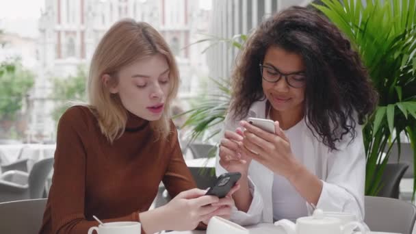 Deux jeunes belles amies assises dans un café à l'extérieur, tout le monde regarde son smartphone. Deux jolies femmes utilisent les médias sociaux sur smartphone dans un café, génération moderne — Video