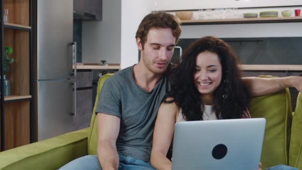 快乐的年轻夫妇坐在沙发上悠闲自在地享受着使用笔记本电脑的乐趣。恋爱中的男女坐在客厅舒适的沙发上，一边看笔记本电脑，一边聊天，一边调情. — 图库视频影像