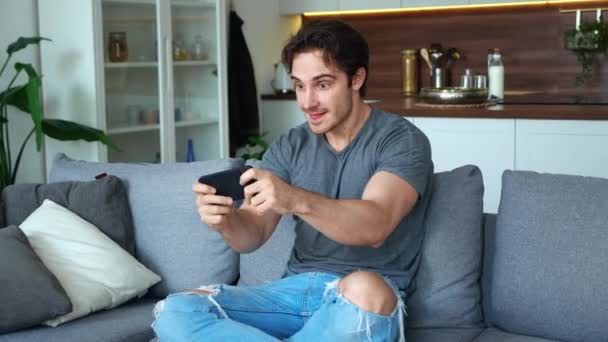 Joven jugando en su teléfono inteligente emocionante aplicación móvil ganar un juego de carreras apretando puño regocijo mientras se sienta en el sofá en la sala de estar en casa. — Vídeo de stock