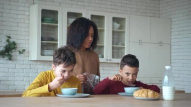 Kahvaltının tadını çıkaran, çikolatalı gevrek yiyen, mutfak masasında oturan mutlu sevimli çocuklar. Güzel Afrikalı Amerikalı anne, tapılası oğullarına hayranlık duyuyor. Sabah yemeğinde onlara nazikçe bakıyor.