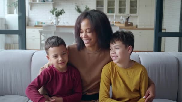 Bella donna di etnia afroamericana che abbraccia delicatamente i suoi due adorabili figli seduti accanto sul divano, ridendo, sorridendo guardando la fotocamera sullo sfondo della cucina di casa. Felice concetto di maternità — Video Stock