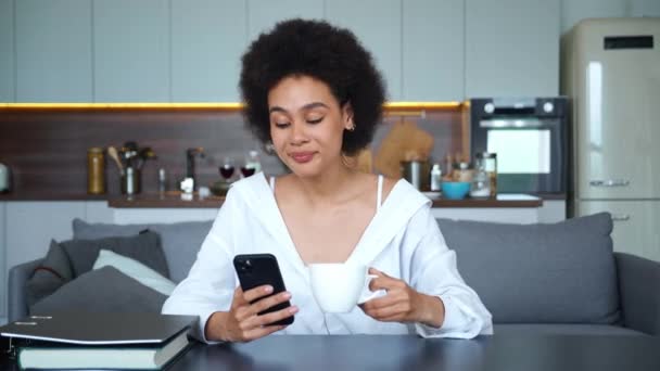 Retrato seguro de una hermosa joven mestiza afroamericana vestida con ropa casual con una taza de café y un teléfono móvil en las manos, descansando sentada a la mesa en el fondo de la cocina — Vídeos de Stock