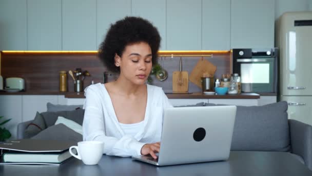 Joven mujer afroamericana seria que trabaja en el ordenador portátil, mirando atentamente a la pantalla de la computadora mientras escribe el texto, sentado en la mesa en la cocina en casa. Joven freelancer trabajando remotamente — Vídeos de Stock