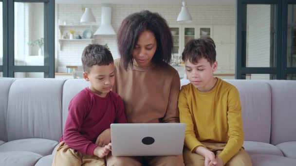 Serene Afro-Amerikaanse etniciteit jonge vrouw geconcentreerd op het werken op afstand op laptop, het typen van tekst, surfen op internet. Vrolijke kinderen leunen op hun moeder, kijken naar het computerscherm. — Stockvideo