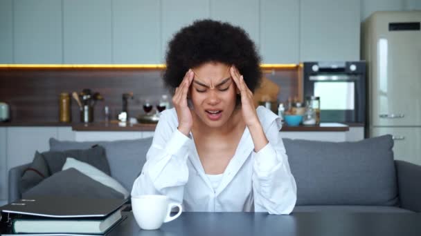 Mujer hermosa afroamericana sentada a la mesa sobre el fondo de la cocina, mirando a la cámara, de repente sintió un fuerte dolor de cabeza, migraña, tomándose de la mano cerca de la cabeza, masajeando sus sienes. — Vídeos de Stock