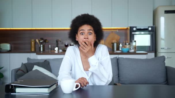 Mujer afroamericana mirando directamente a la cámara del monitor, expresando emociones de horror al recibir malas noticias, cubriéndose la boca y sosteniendo sus sienes con la mano, sentada en la mesa de su casa — Vídeos de Stock