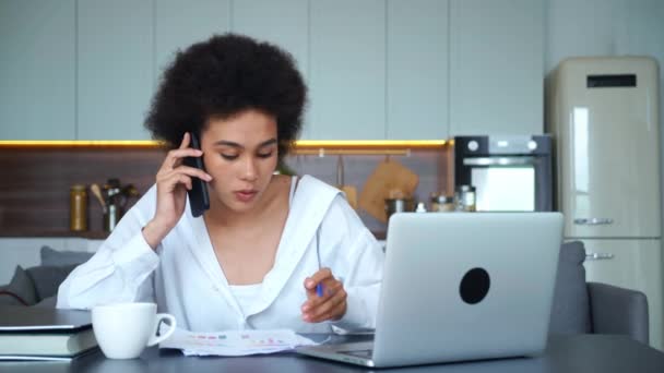 Молодая красивая афроамериканка работает удаленно из дома на ноутбуке с графиками, разговаривает по мобильному телефону, проверяет информацию на бумаге с содержанием на экране монитора, делает заметки с ручкой. — стоковое видео