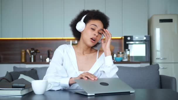 Jovem afro-americana bonita mulher em fones de ouvido sem fio se sente exausta, fecha seu laptop e se inclina na parte de trás do sofá. Uma mulher freelance ocupado, estudante cansado após o e-learning ou trabalho remoto — Vídeo de Stock